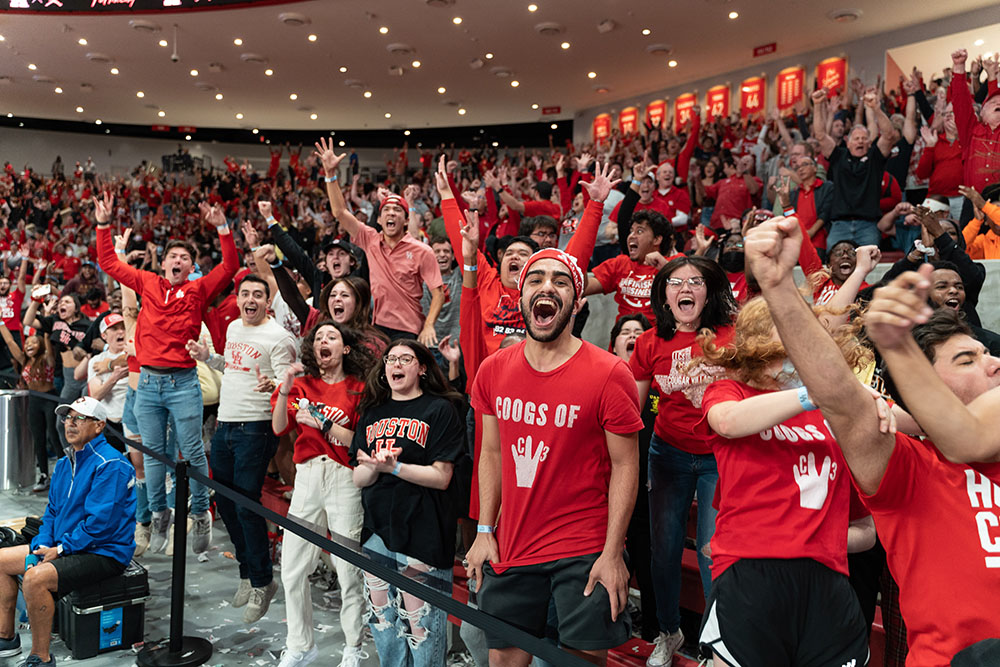 A large group of students in the Fertitta Center jump off the ground in excitement as they cheer for the UH men's basketball team.