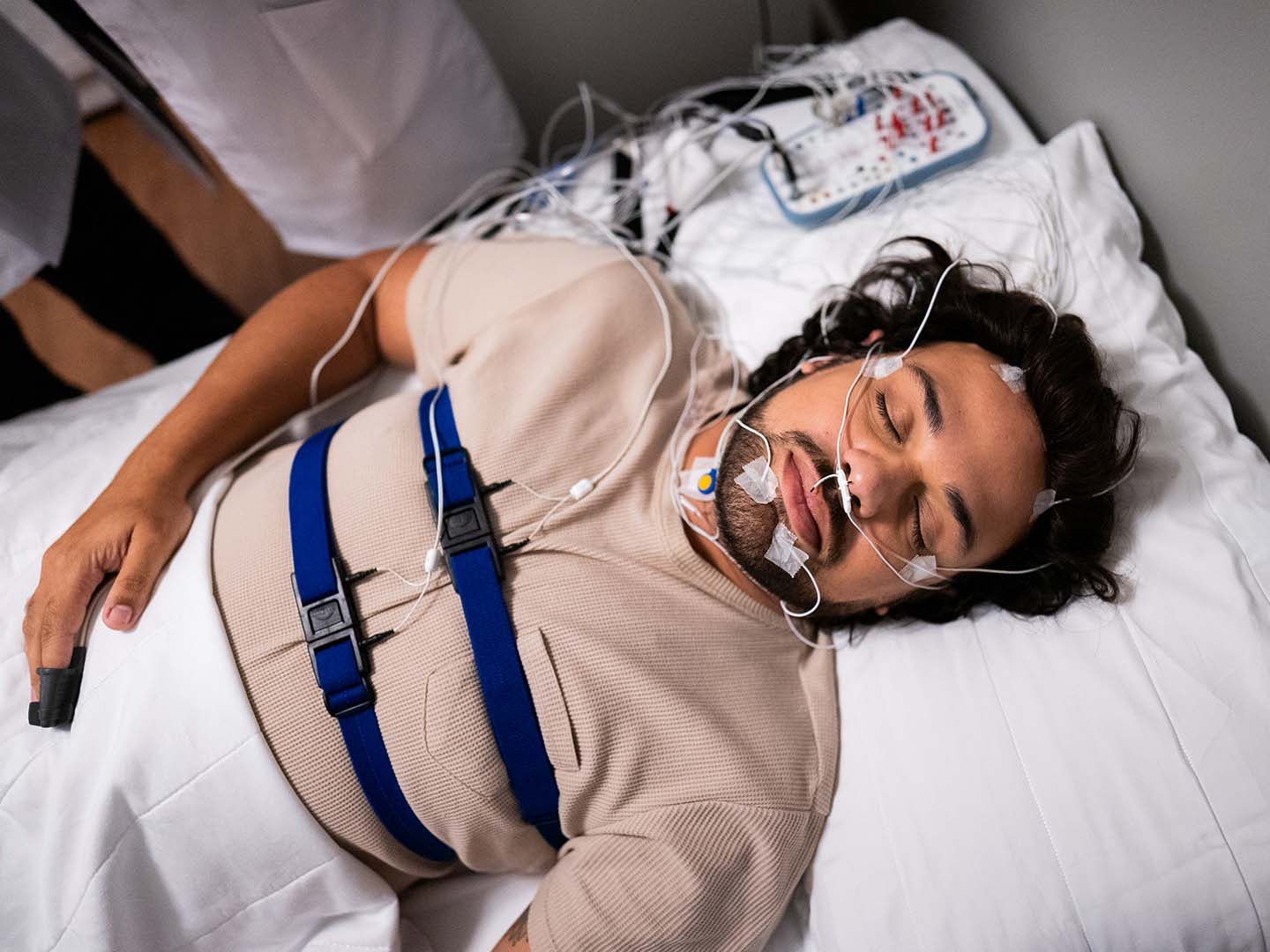 A person is undergoing a sleep study, connected to various sensors and equipment designed to monitor physiological functions while lying in a medical bed.