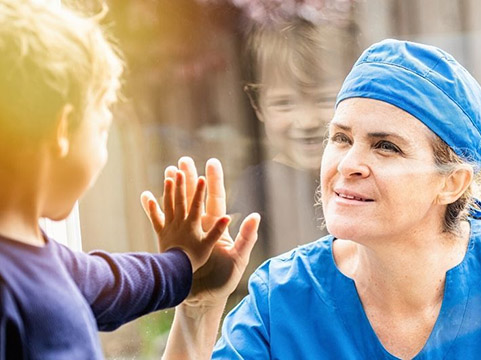 A woman in blue healthcare scrubs touches a glass window. A young boy on the other side of the window places his hand on the woman's hand.