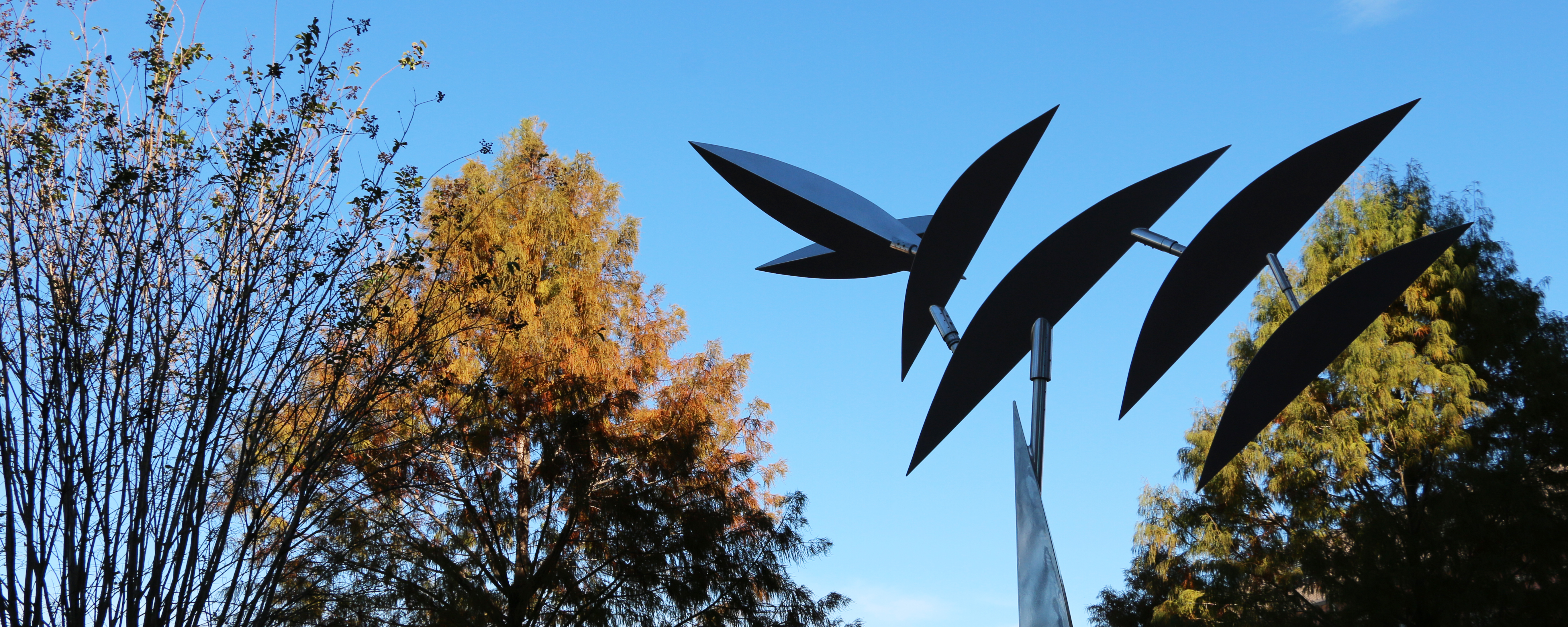A metalic sculpture with leaf-like parts in the foreground and trees with red and green leaves in the background