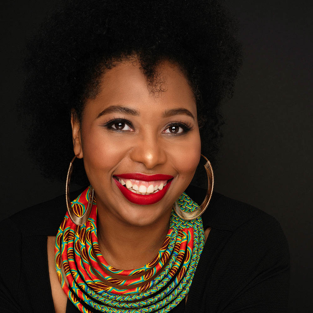 Portrait of a black woman smiling. She wears gold hoop earrings and a large multi-colored circular necklace.