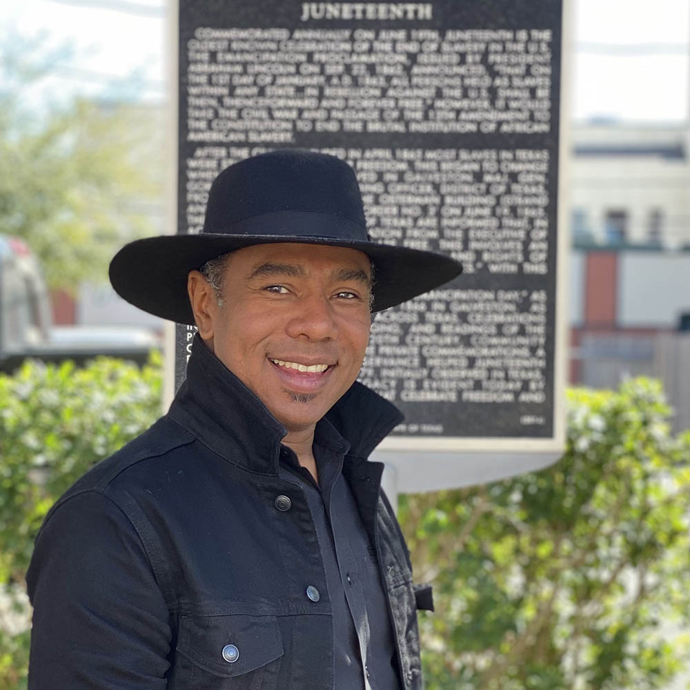 A black man smiles wearing a black wide-brimmed hat and black dress shirt. In the background is a large silver and black plaque commemorating Juneteenth.