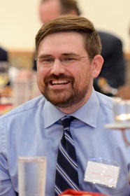 A portrait of a smiling man with a beard wearing a blue dress shirt and tie.