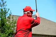 A grey-haired man wearing red swinging a golf club 
