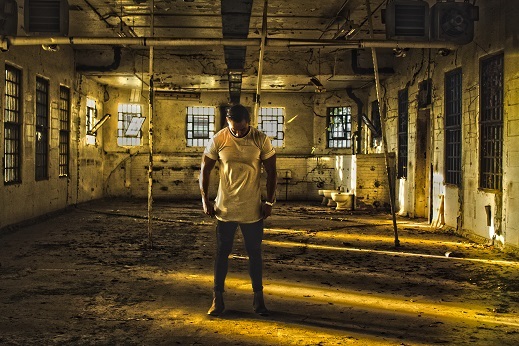 A lone figure in yellow clothes looking down standing in a dilapidated room, bathed in yellow light
