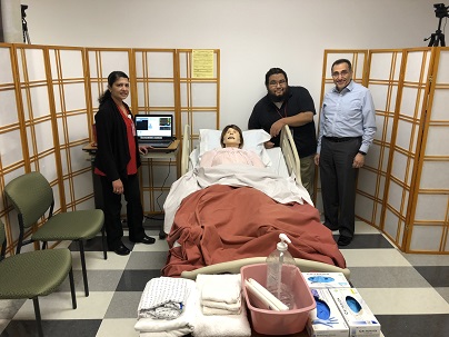 Three people standing next to a simulation manikin laying down on a  hospital bed