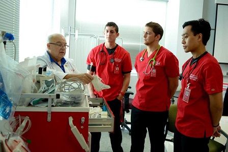 Three male students in red scrubs listen to a bald male instructor in a labcoat present equipment in red drawer