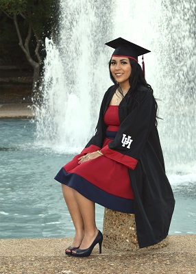 A raven-haired hispanic woman wearing black graduation regalia sitting in front of a fountain