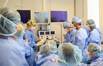 A group of people wearing surgical scrubs and protective wear huddled around a piece of equipment with screens