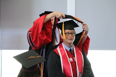 A woman in red and black academic robes places an academic hood over a man in glasses sitting