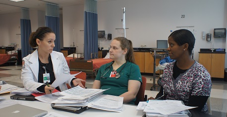 A female instructor in a labcoat sits next to two female students at a table with binders and notes