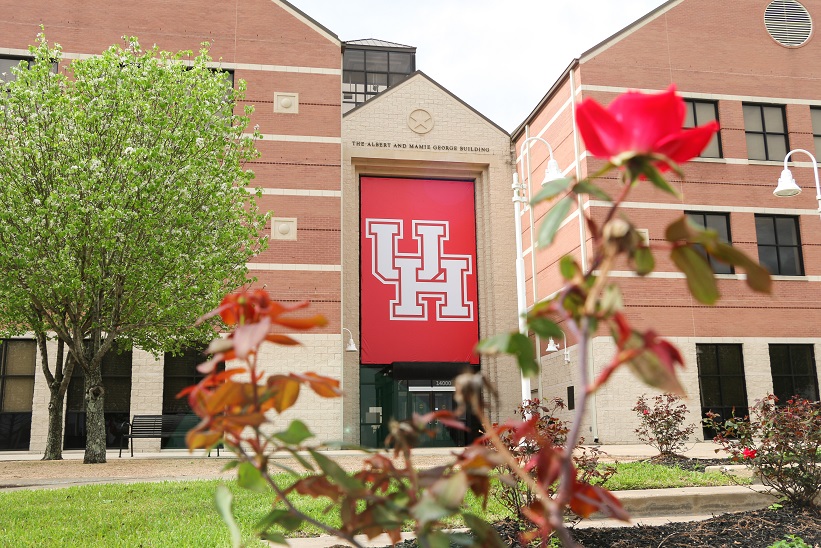 The front entrance of a three story brick building. A red banner with the UH logo covers the front side, and red flower is in the foreground