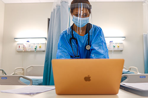 A nurse wearing personal protective equipment standing at table while working on a laptop