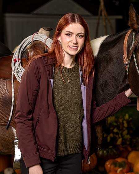 Portrait of a red-haired woman smiling and standing next to a brown horse.
