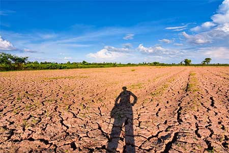 The silhouette of a farmer stands facing a large field of dry cracked land.