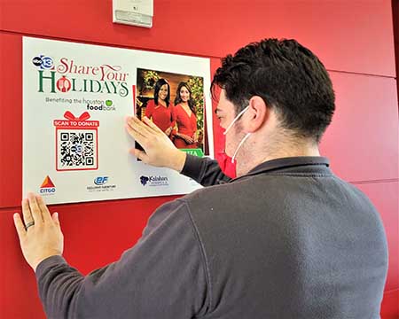 A man hangs a large poster on a red wall. The poster advertises the Share Your Holidays food drive.