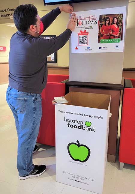 A man hangs a poster on a pillar in a lobby. The poster advertises the Share Your Holidays food drive.