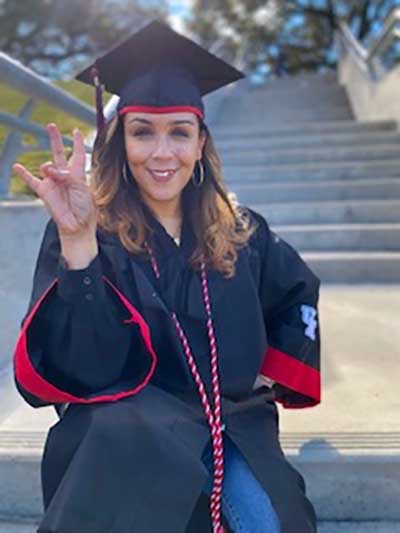 Photo of a hispanic woman smiling and wearing graduation regalia. She holds her right hand up in the "cougar sign" gesture: her ring finger folded toward her palm.