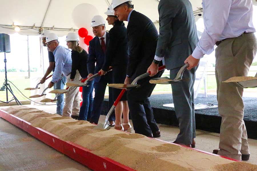 A group of people in suits and hardhats lift shovels with dirt in unison