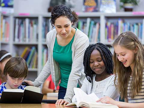 A group of young students read books in a library. An adult stands at the table between two students.