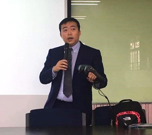 An Asian man in a suit speaks in a microphone while holding a virtual reality headset in his hand.