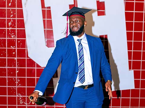 A black man wearing a blue suit and a black graduation cap opens a bottle of champagne in front of a white University of Houston logo on a red tile wall.