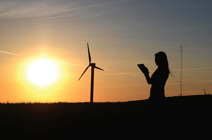 A silhouette of a woman in a field at sunset. The silhouette of a wind turbine is visible on the horizon.