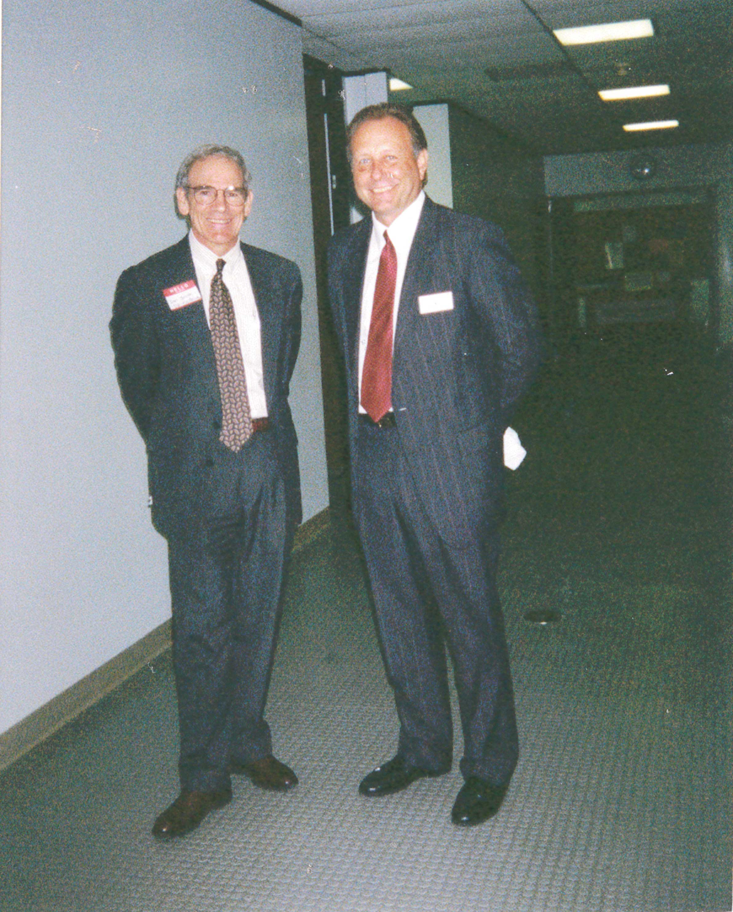 Photograph of two men in suits standing in a hallway.